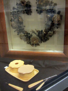 Color photo of An 1880s ornate hair wreath along with a hair receiver and tools for collecting hair for the art.