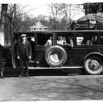 Black and white photograph of two men standing in front of a long car with men inside the car.