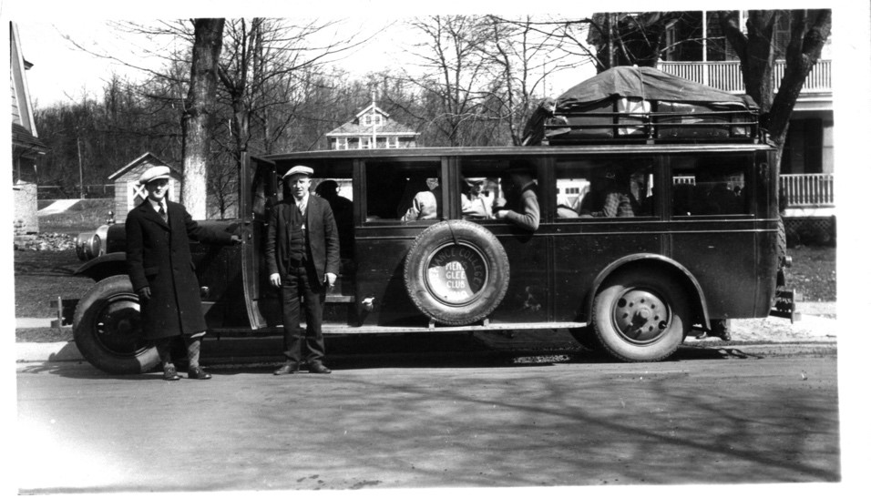 Black and white photograph of two men standing in front of a long car with men inside the car.