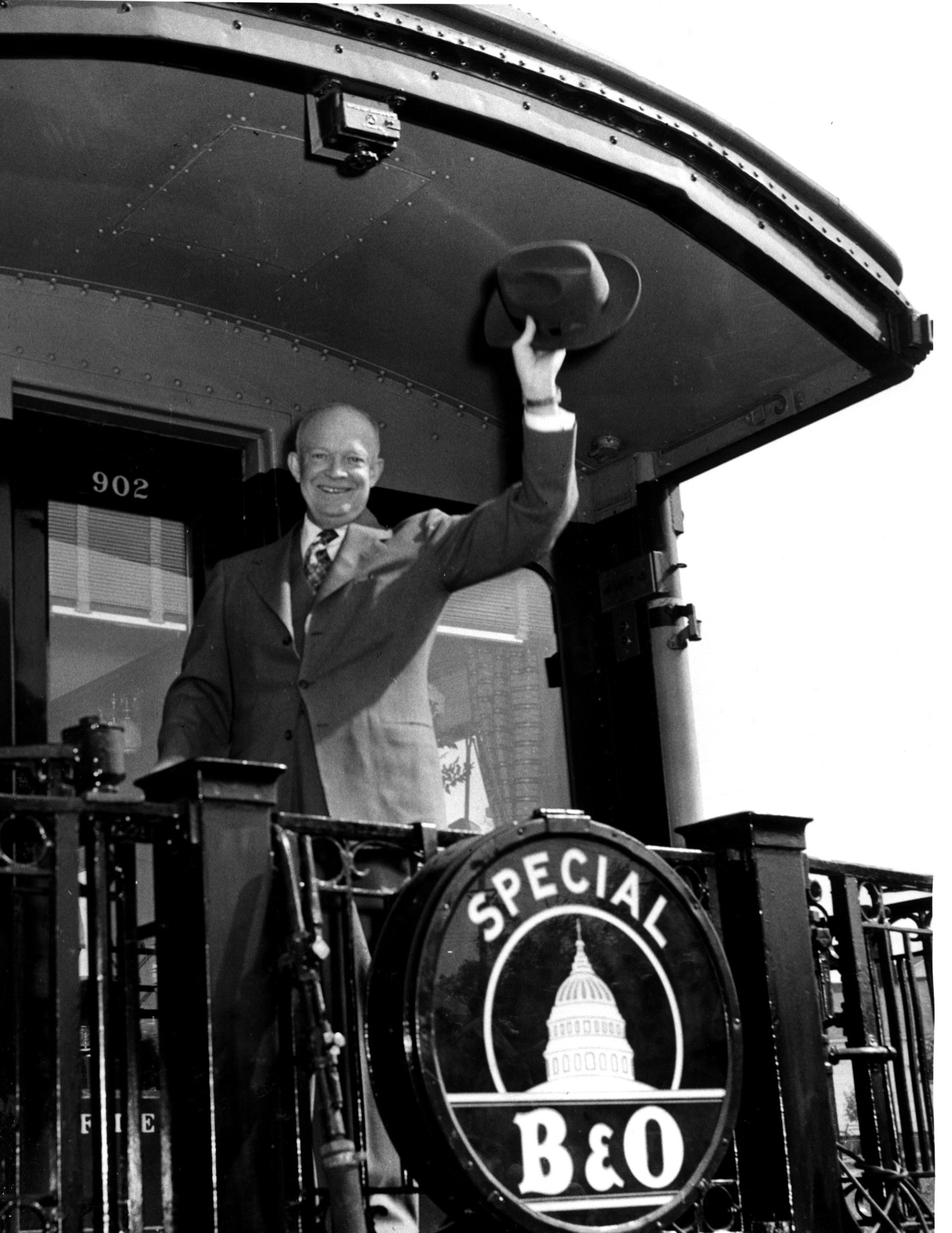 Black and white photograph of a man standing on a train platform and waving his hat.