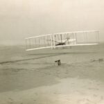 Black and white photograph of an airplane on a beach. A man stands on the right facing the airplane.