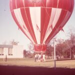 A red and white hot air balloon inflated with the basket on the ground. A group of people standing by the balloon's basket.
