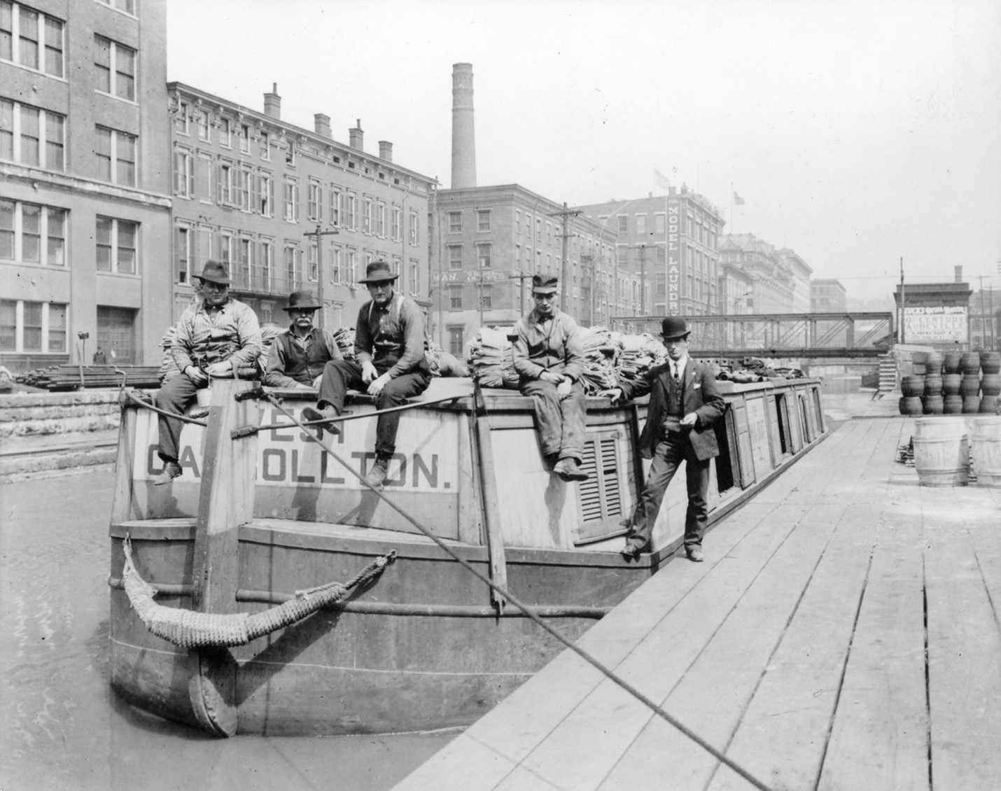 Black and white photograph of five men standing on a canal boat. Building line the canal in the background.
