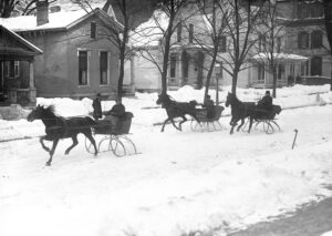 Black and white photograph. Three sleighs pulled by horses on a snowy road.