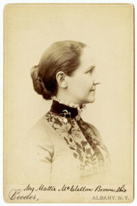 A historic photograph in sepia tones of a woman in profile. She is wearing a high-collared dress and has her hair in a bun.