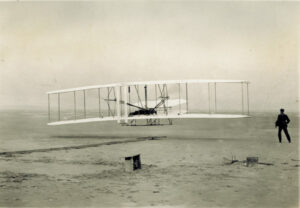 Historical image in sepia tone. Shows an airplane on a beach and a man in dark clothing standing on the right side.