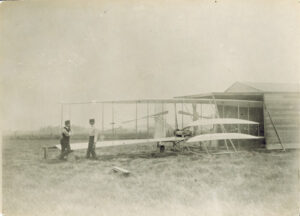 A historic photograph in sepia tone of an airplane with two men standing on the ground by the left airplane wing.