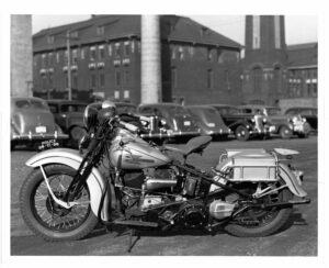 A black and white historical photograph of a Harly Davidson police motorcycle. It is parked in front of a brick building.