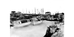 A black and white historic photograph of canal boats in the water in a canal. A house is in the background.