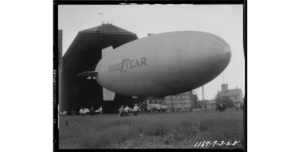 A black and white historic photograph of a large blimp with Goodyear printed on the side. A large hanger is in the background.