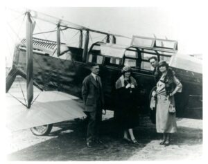 A historic black and white photograph of four people standing in front of a small airplane.