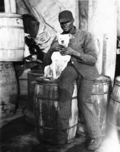 Man wearing cap and attire of a boat crew member smiles while seated on a barrel with his hands gripping a white dog at his side