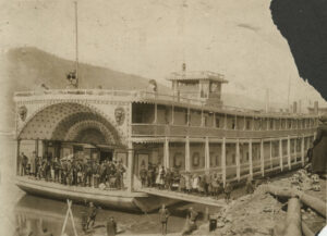 Sepia toned photo of Cotton Blossom showboat on the banks of the Ohio River with band members on board and children posing on the shore and boat ramp