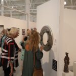 A photograph of a group of people looking at an art exhibit. There is a round piece of art hanging on a white wall.