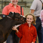 Photograph of a girl wearing a red jacket petting a brown cow. A woman wearing a pink shirt stands behind the cow.