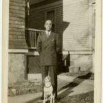 Black and white photograph of a man standing in front of a house. A dog stands at his feet.