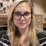 Portrait of a woman with long blond hair wearing glasses. Background is an office setting.