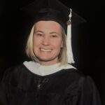 Portrait of a woman in a black cap and gown. She has blond hair and is smiling. Background is solid black.