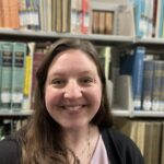 Portrait of a woman with long dark hair smiling. The background is books on library shelves.