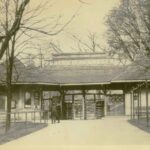 Historical photograph of the entrance of the zoo. A large sidewalk leads to a wooden gate building.
