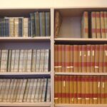 A photograph of six book shelves with many books on them. Most of the books have brown and red spines.