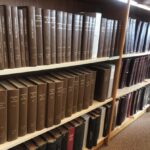 A photograph of book shelves with three rows of books. Most books have brown spines.