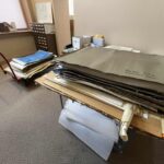 A photograph of a table with large folders and blueprints on top. Another cart with blueprints is in the background.