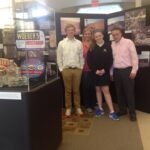 A group of four people standing in front of an exhibit panel. Another exhibit case is on the left side of the photograph.