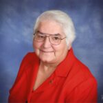 Photograph portrait of a person with short white hair and glasses. She is wearing a bright red shirt.