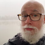 A portrait photograph of a person wearing a black shirt and standing in front of white background. Jim has a beard and is wearing glasses.
