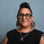 A portrait photograph of a person wearing a black shirt in front of a blue background. Michelle has short dark hair and is wearing glasses, earrings, and a necklace. 