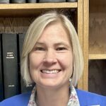 A portrait photograph of a person wearing a blue sweater and flowered shirt and standing in front of a book shelf. She has shorter blonde hair.