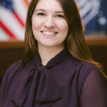A portrait photograph of a person wearing a dark purple shirt. She has long brown hair.