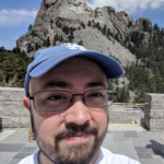 A portrait photograph of a person wearing a white shirt and blue hat and standing in front of a mountain. Kris is wearing glasses and has a beard.