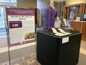 Photograph of an exhibit. Left side of the photograph shows a poster with text. Right side of the image shows a display case with a purple shirt and some papers in front.
